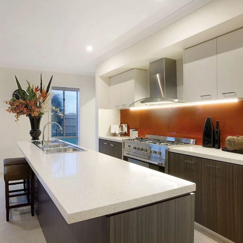 Modern kitchen with a long white island countertop, brown wooden cabinets, and an orange backsplash. A stovetop with a metallic hood is visible. A vase with tall flowers decorates the counter. A window provides natural light to the space.