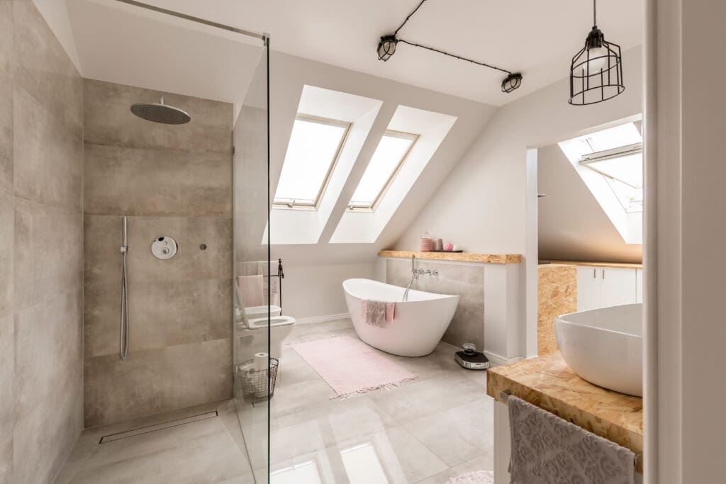 Modern bathroom with slanted ceiling and skylights. Features a glass-enclosed shower, freestanding bathtub, and vessel sink on a wooden counter. Neutral tiles and soft lighting create a serene ambiance. Pink accents add a touch of color.