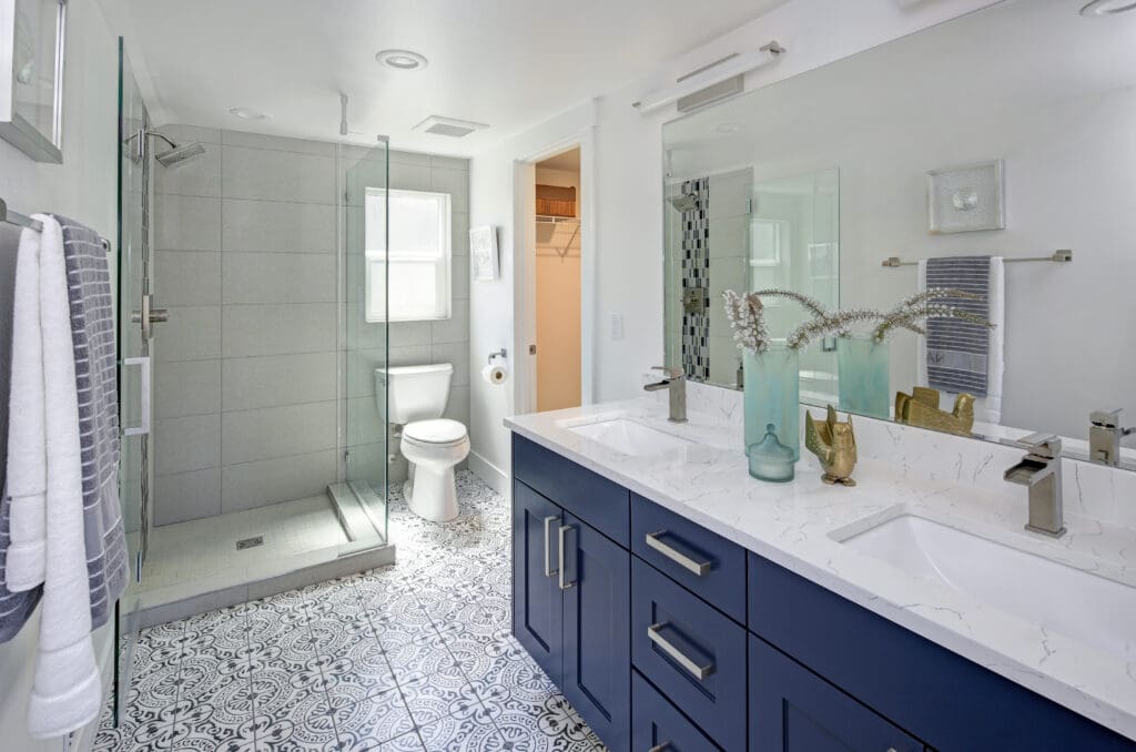 Modern bathroom with blue double-sink vanity, white countertop, and large mirror. A glass-enclosed shower and toilet sit on patterned floor tiles. Towels hang on the wall, and decorative vases adorn the counter.
