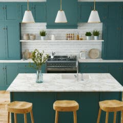 Modern kitchen with teal cabinets, white marble countertops, and wooden barstools. Three pendant lights hang above the central island. Green plants and kitchenware decorate the space, with a brick backsplash and a central stove. Natural wood flooring.