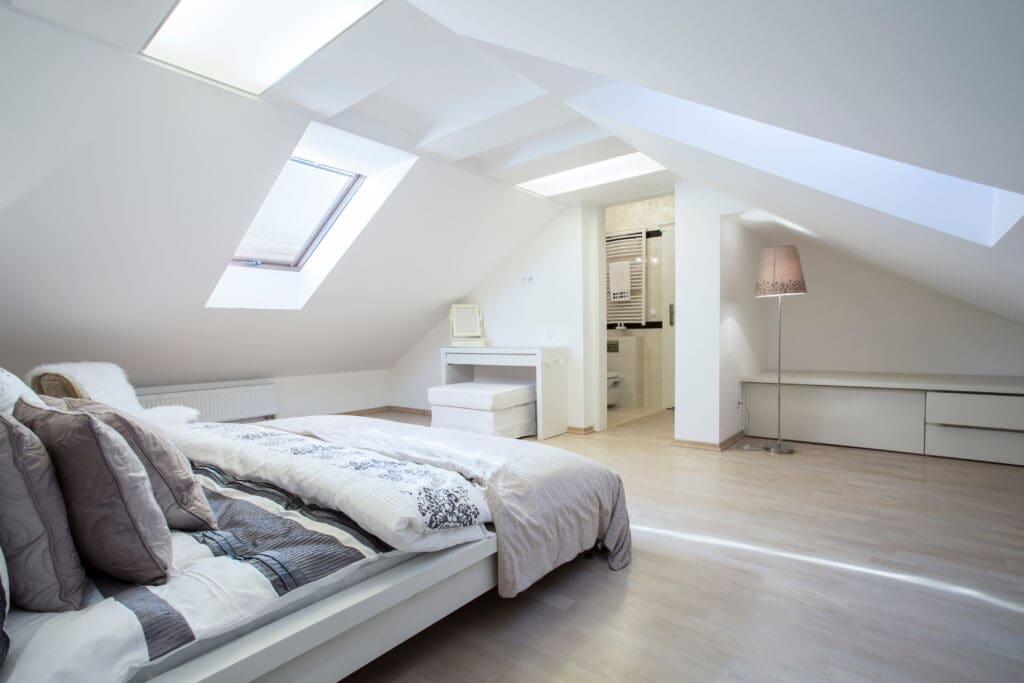 Bright attic bedroom with a sloped ceiling, featuring a bed with patterned pillows, a skylight, and wooden flooring. A small vanity with a mirror is near the bathroom entrance. A floor lamp stands beside low cabinets along one wall.