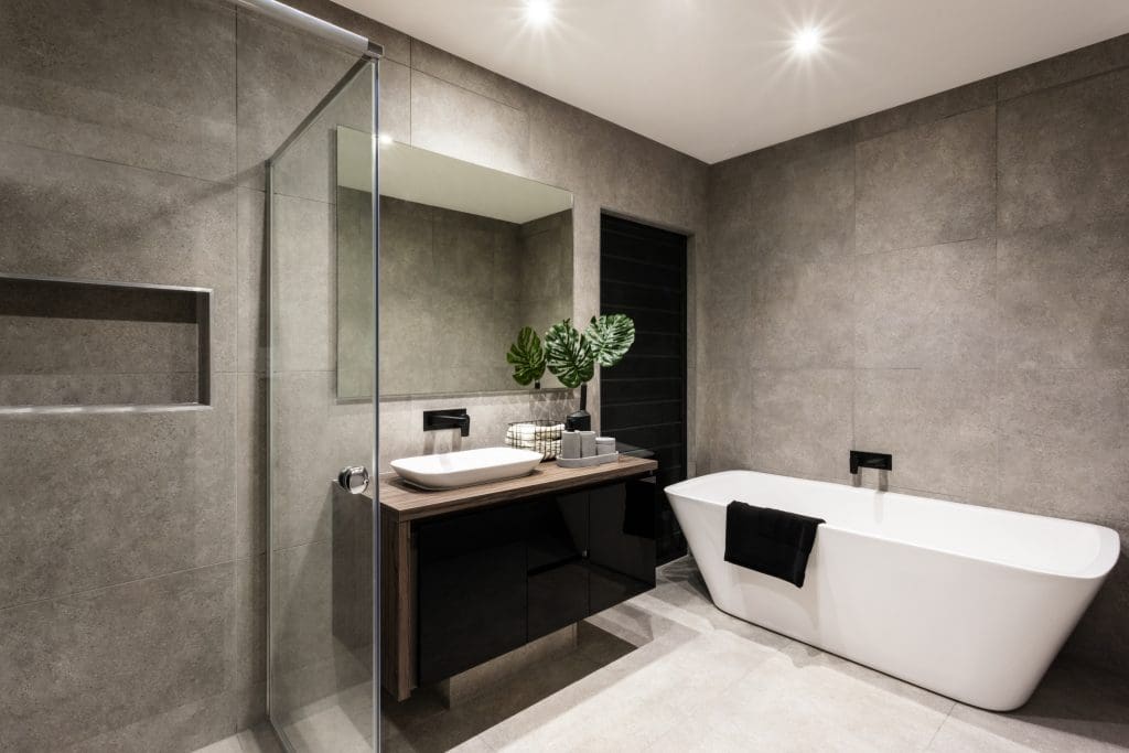 Modern bathroom with a glass shower, freestanding white bathtub, and a vanity with a vessel sink. Gray tiled walls and floors create a minimalist look. A large mirror, plants, and neatly arranged toiletries complete the decor.