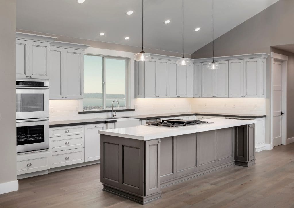 A modern kitchen featuring white cabinets, a large island with a gray base and white countertop, stainless steel appliances, a double oven, pendant lights, and a window with a view. The floor is light wood, and the walls are painted light gray.