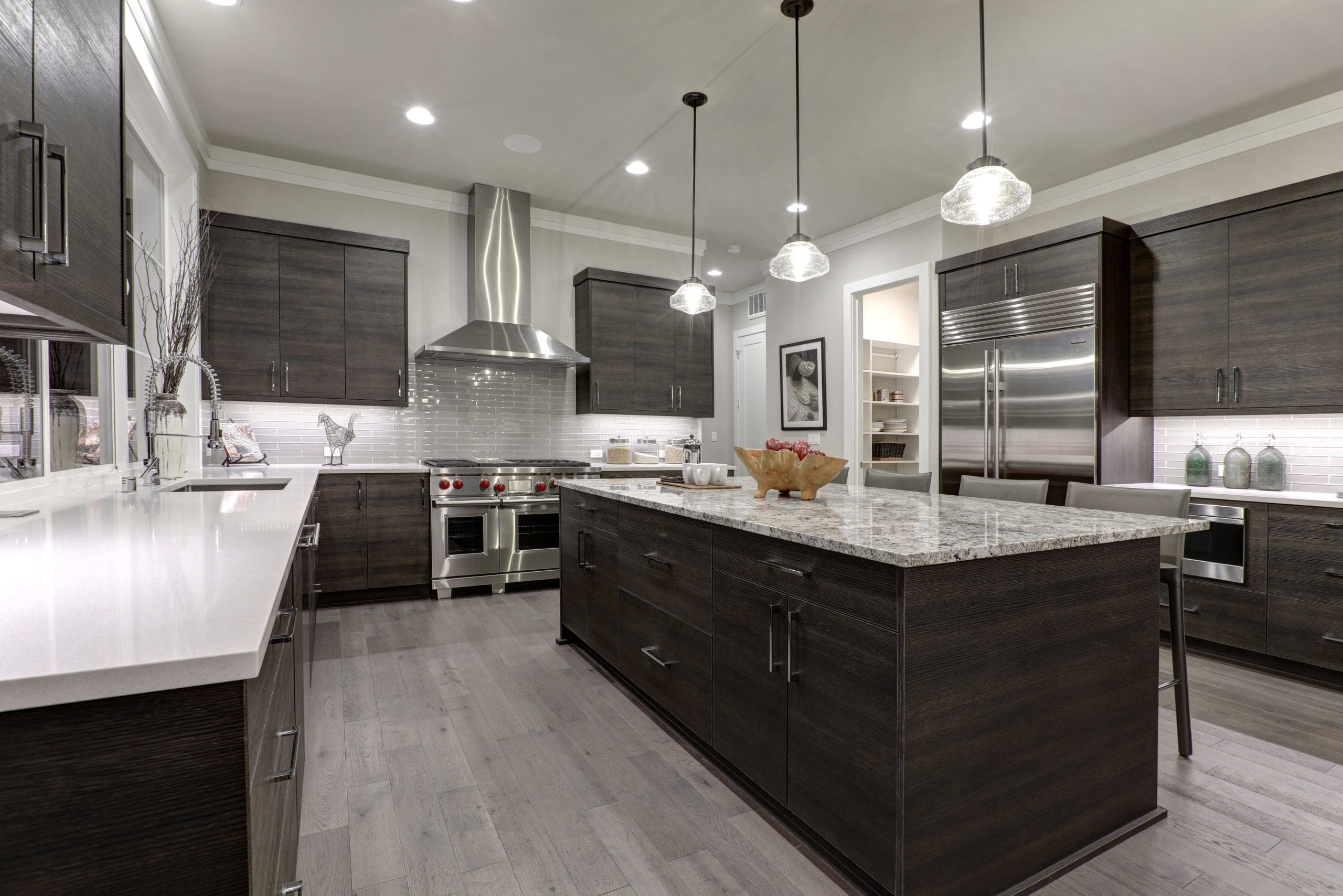 Modern kitchen with dark wood cabinets, stainless steel appliances, an island with granite countertop, pendant lights, and light wood flooring. A backsplash of light-colored tiles and decor accents complete the sleek design.