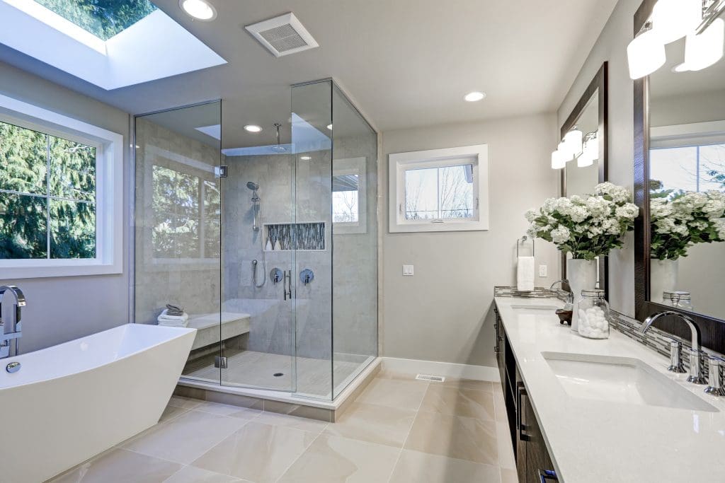 Modern bathroom with a freestanding bathtub and glass-enclosed shower on a tiled floor. A double sink vanity with a large mirror is adorned with a bouquet of white flowers. Natural light filters through a window and skylight.