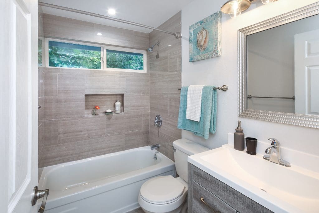 Modern bathroom with a white bathtub and shower, gray tile walls, and a small window. A vanity with a mirror, sink, and toiletries is on the right. Towels are hung on the wall, and a decorative art piece is above the toilet.