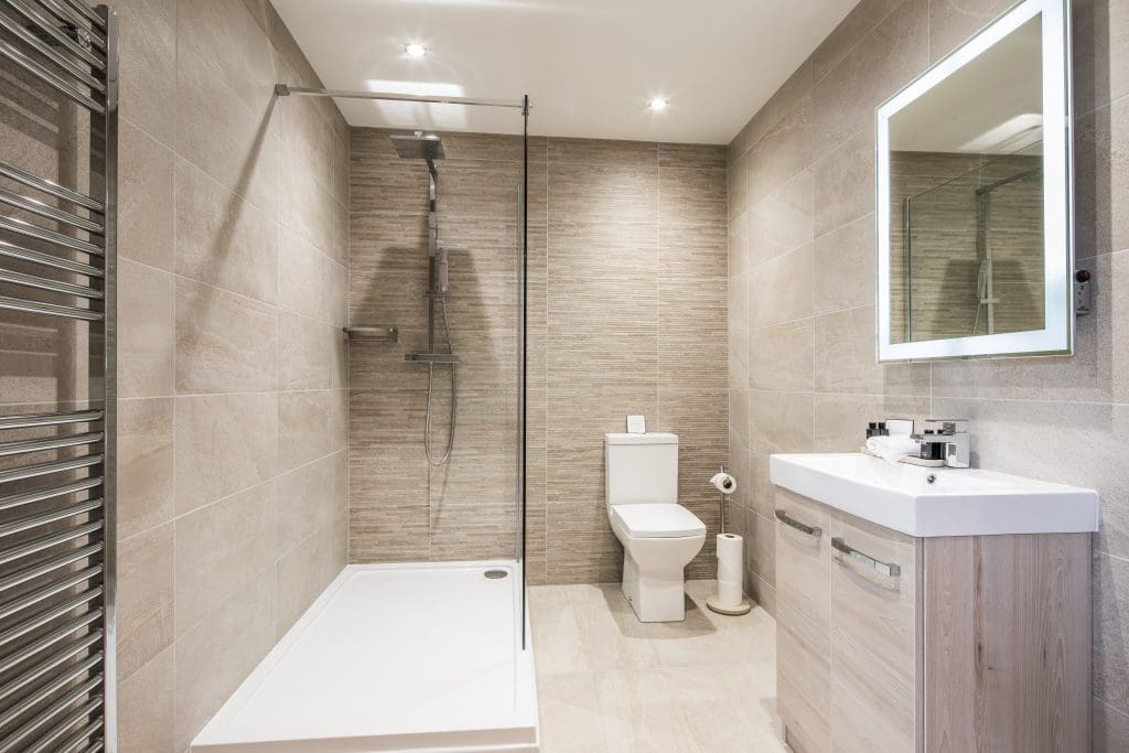 Modern bathroom with a walk-in shower, glass partition, and silver fixtures. Beige tiled walls and floor. A white sink with a mirror and toiletries on the right. Toilet with a roll holder. Ceiling lights illuminate the space.
