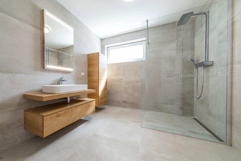 Modern bathroom with large gray tiles, a walk-in glass shower, and a wall-mounted rain showerhead. A wooden floating vanity with a rectangular sink and illuminated mirror is on the left. A small window provides natural light.