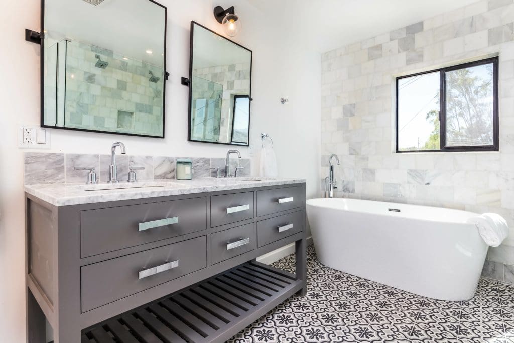 Modern bathroom featuring a double sink vanity with a marble countertop, large mirrors, and a light fixture. A freestanding bathtub is positioned near a window. The floor is covered with patterned tiles, and the walls have light-colored tiles.