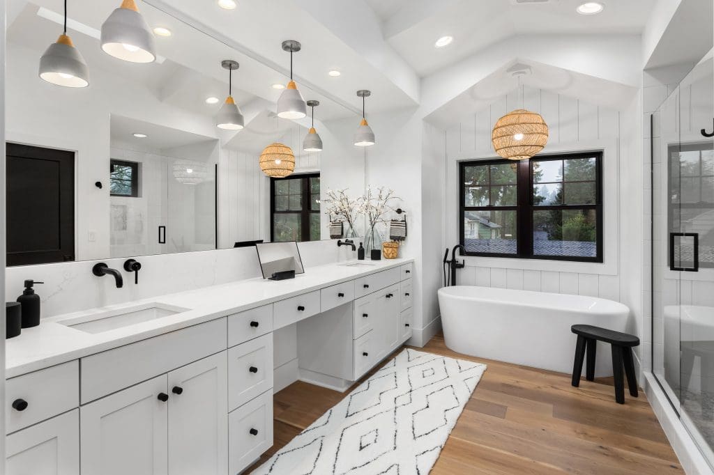A modern white bathroom with a double vanity, large mirrors, and black fixtures. It features pendant lights, a freestanding bathtub, a glass shower, and a patterned rug on wooden floors, with windows providing natural light.