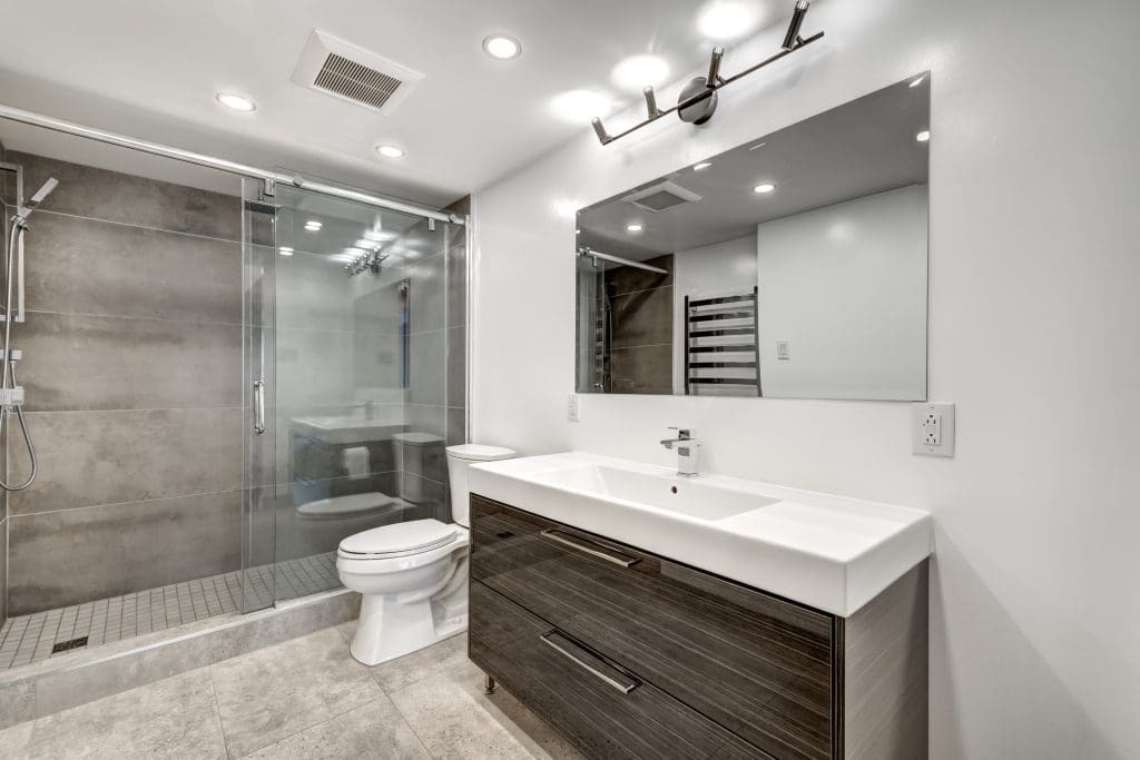 Modern bathroom with a large mirror above a dark wood vanity and white sink. A toilet is next to a glass-enclosed shower with a rain showerhead. The room features recessed ceiling lights and light gray floor tiles.