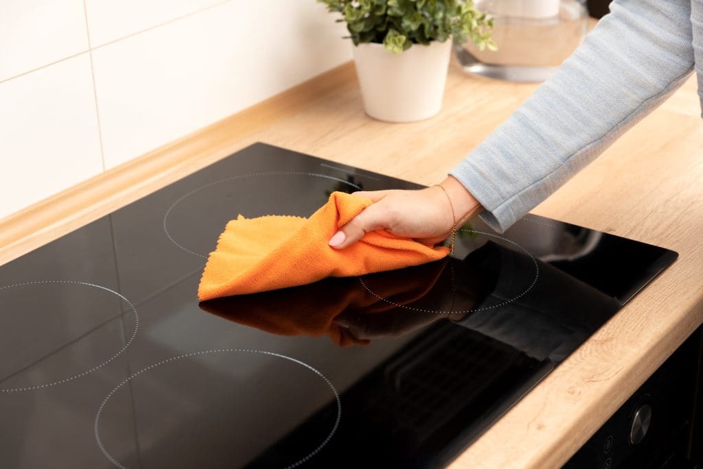 A person cleaning a black glass stovetop with an orange cloth. There is a wooden countertop and a potted plant in the background.