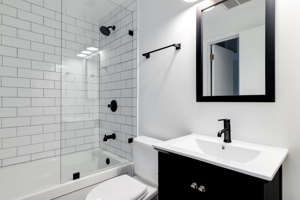 Modern bathroom with a white subway-tiled shower and glass door. Features a white toilet, black fixtures, a white sink with a black vanity, and a black-framed mirror. Bright lighting reflects on the clean, minimalist design.