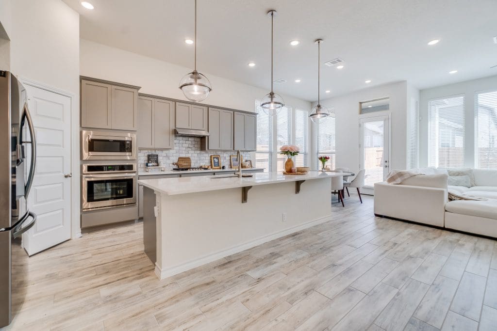 Modern kitchen and living area with light wood flooring. Features an island with seating, pendant lights, stainless steel appliances, light gray cabinets, and a cozy white couch. Large windows provide ample natural light.