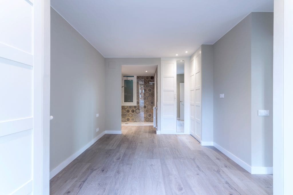 Minimalist interior with light wood flooring, pale walls, and a built-in wardrobe. A doorway leads to a bathroom, visible with a patterned tile wall. The space is bright with recessed lighting and a contemporary design.