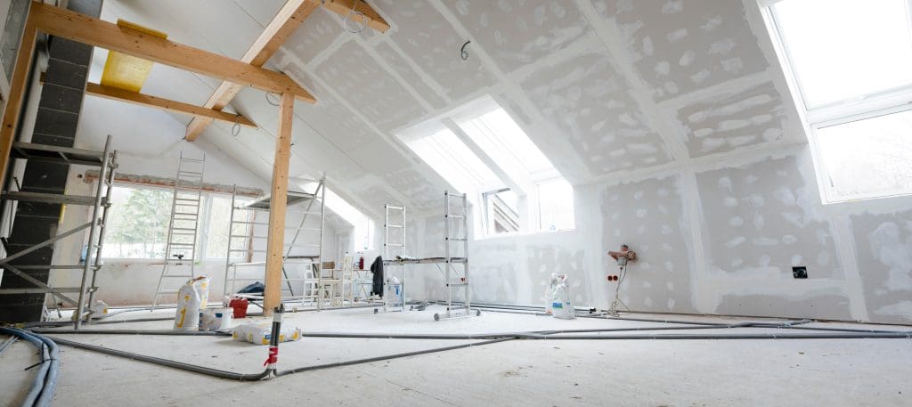 A bright, unfinished attic space under renovation with exposed beams, scaffolding, ladders, and construction materials. The walls are partially covered in drywall with visible seams, and light streams in through large skylights.