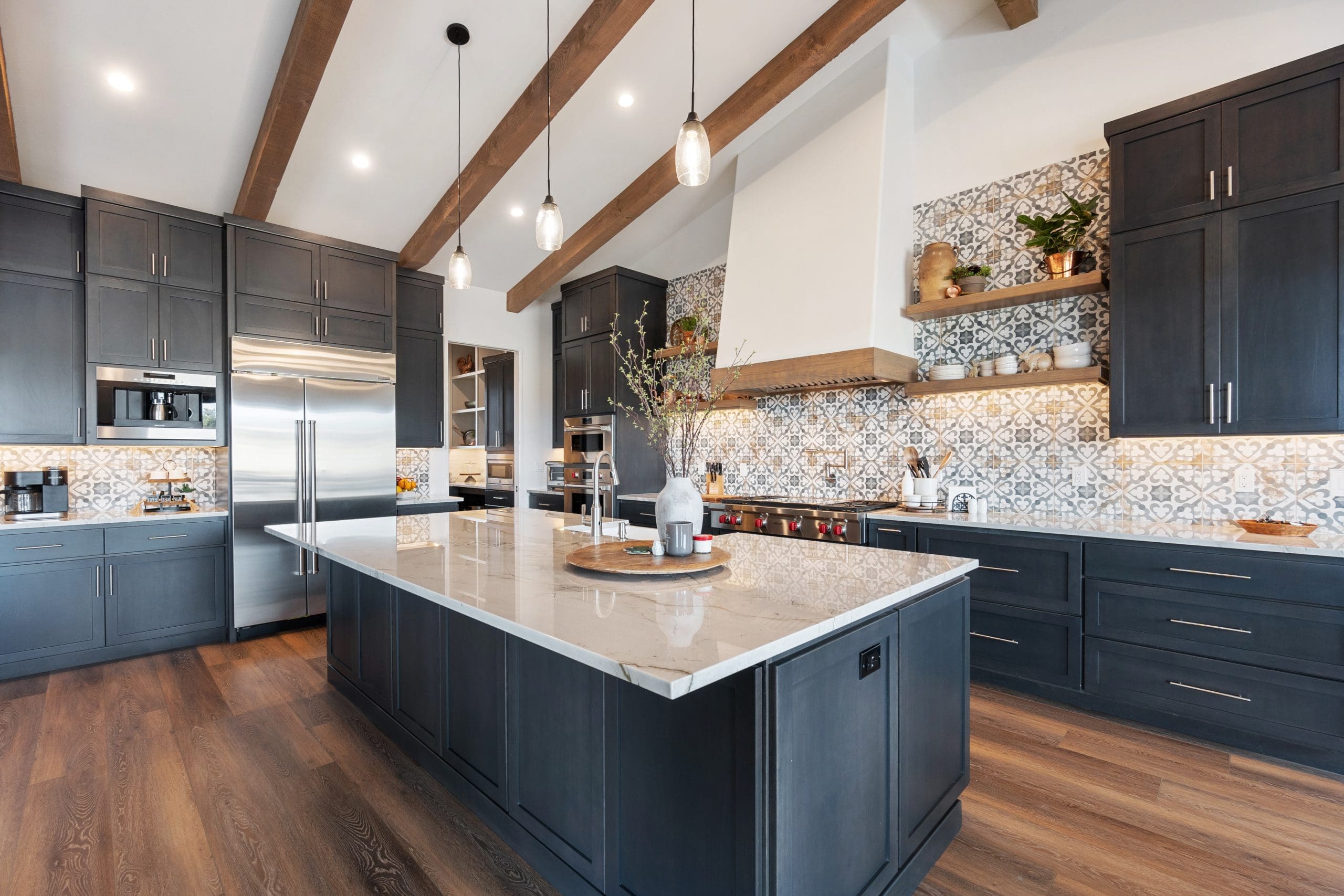 A modern kitchen featuring dark wood cabinets, ornate patterned backsplash, and an island with a light marble countertop. Pendant lights hang from the ceiling with wooden beams. Stainless steel appliances and decorative plants add touches of elegance.