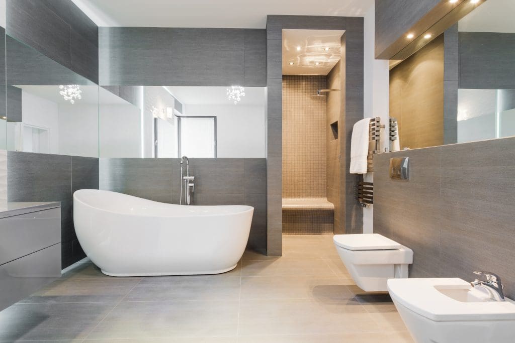 Modern bathroom with a white freestanding bathtub, wall-mounted toilet and bidet, walk-in shower with bench, large grey tiles, and illuminated mirrors. Neutral color palette creates a sleek, contemporary aesthetic.