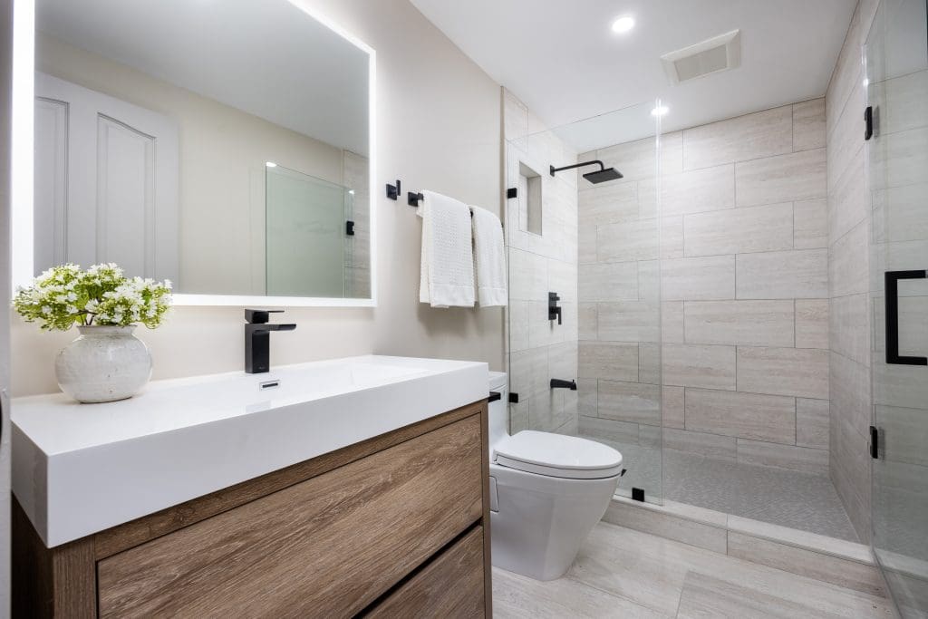 Modern bathroom with a sleek design. Features a wooden vanity with a white countertop, rectangular mirror with backlighting, and black fixtures. There's a glass-enclosed shower with light beige tiles and a white toilet. A potted plant adds greenery.