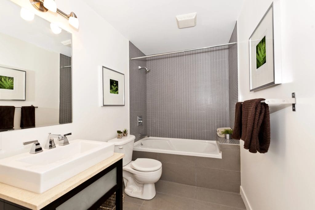 Modern bathroom with a white sink, toilet, and bathtub. The walls are light gray with a tiled accent behind the tub. There are framed pictures on the walls and brown towels hanging. Bright lighting illuminates the space.