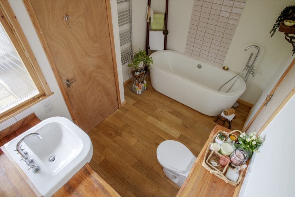 A bathroom with a wooden floor features a white bathtub, a sink with a wooden countertop, and a toilet. A wicker basket with towels and toiletries sits on a shelf. A small plant and various bath items decorate the space.