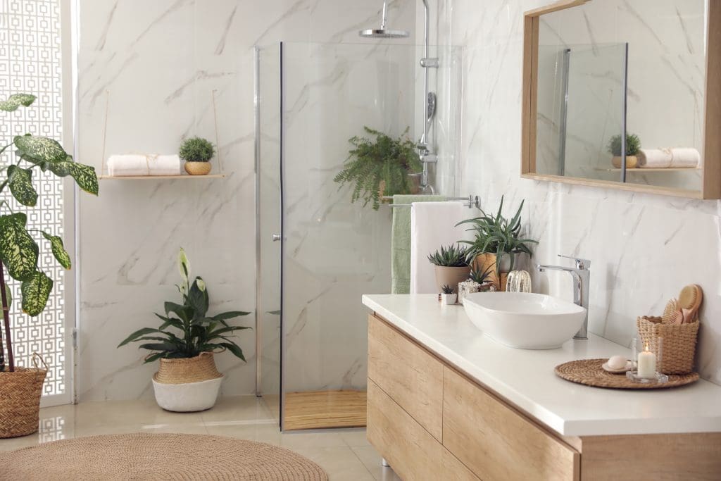 Modern bathroom with a glass shower, wooden vanity, and white sink. Plants decorate the space, along with candles and towels. A large mirror hangs above the vanity. A woven mat is on the floor. Natural light streams in, creating a serene atmosphere.