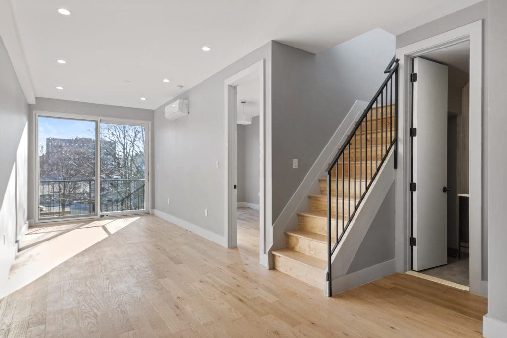 Bright, empty living space with light wood flooring, a staircase with black railings leading upstairs, and a large window and glass door opening to a balcony with a view of trees and buildings. Walls are light gray with recessed lighting.