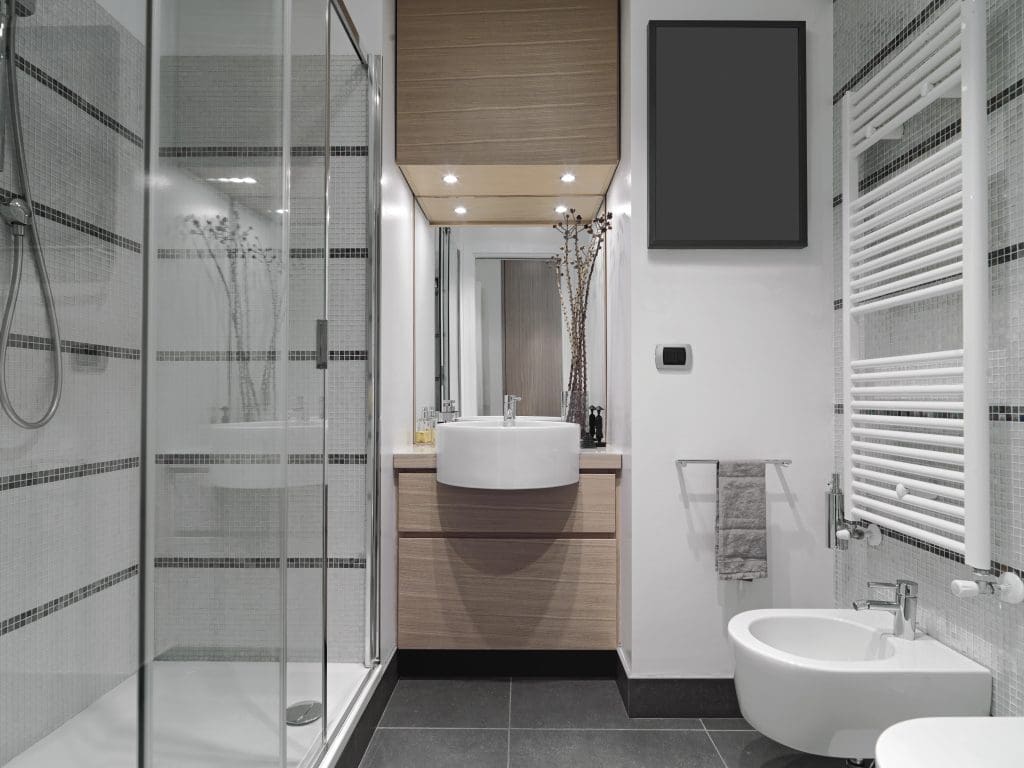 Modern bathroom with a glass-enclosed shower on the left, a wooden vanity with a round white sink and mirror in the center, and a bidet and towel radiator on the right. The decor features neutral tones and a plant arrangement.