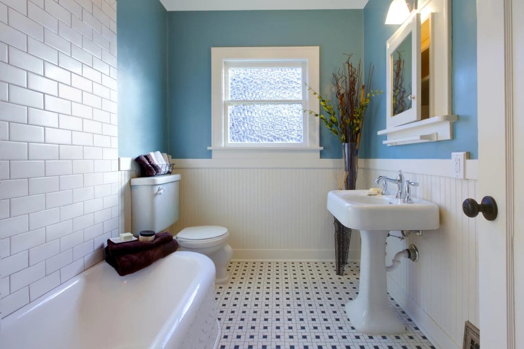 A bathroom with light blue walls features a white pedestal sink, a toilet, and a bathtub with towels. The floor has a white and black checkered pattern. A window above the toilet and decorative plants add to the decor.
