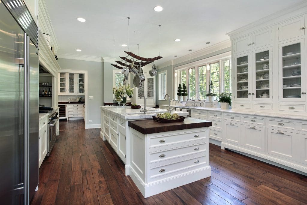 A spacious kitchen with dark hardwood floors and white cabinetry. It features a large island with dark countertops, pendant lights, and hanging utensils. Large windows provide natural light. Stainless steel appliances are visible in the background.