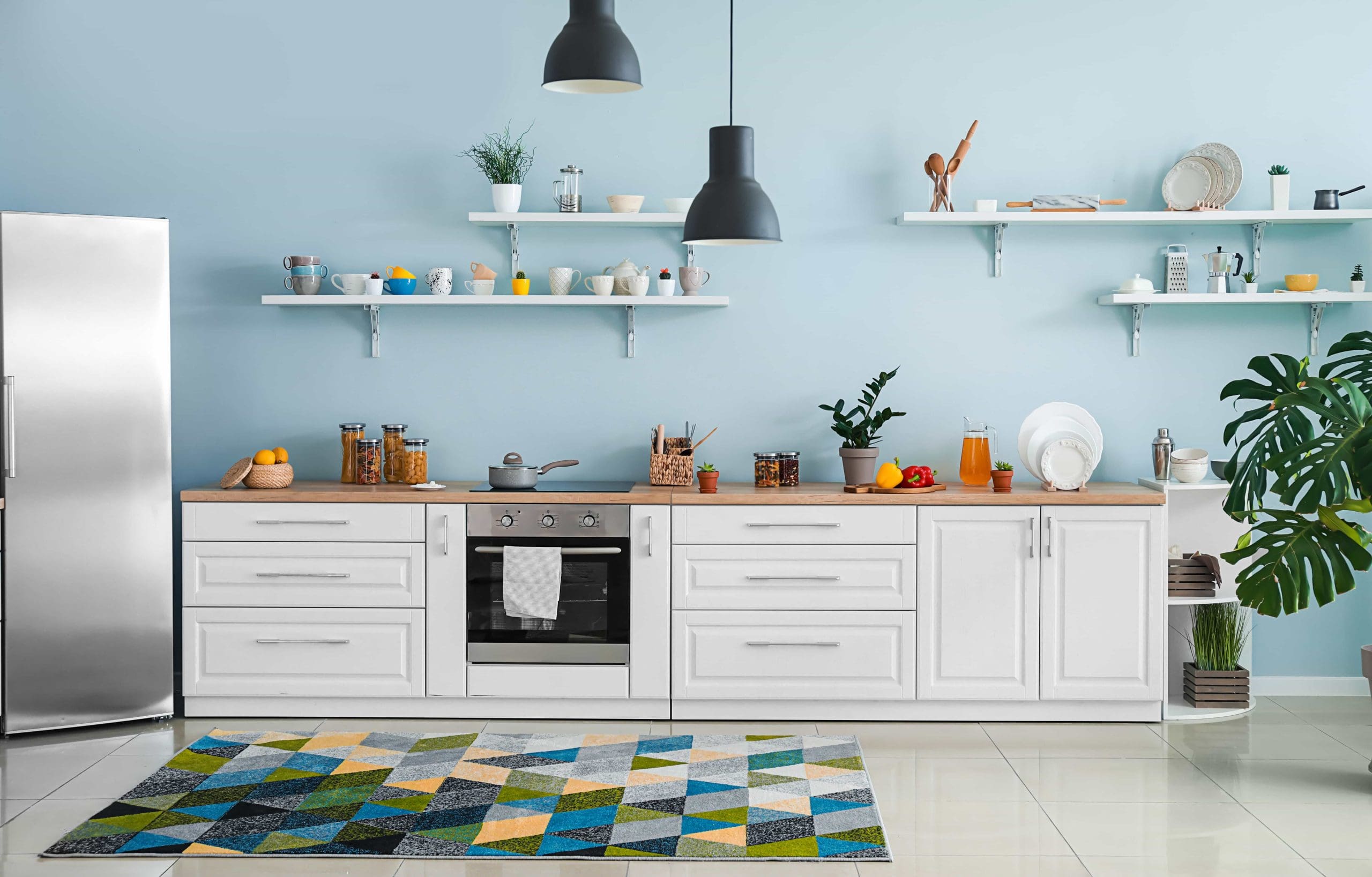 Modern kitchen with light blue walls, white cabinets, and stainless steel appliances. Shelves hold various jars, dishes, and decor. A geometric-patterned rug is on the floor, and leafy plants add greenery. Hanging lamps provide lighting.