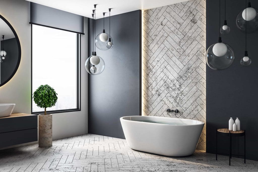 Modern bathroom with a freestanding oval bathtub against a herringbone-tiled wall. Large window with natural light, round mirror, and globe pendant lights. Potted plant on pedestal and toiletries on a round side table.