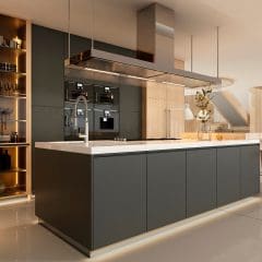 Modern kitchen with a large island featuring a built-in sink and stovetop. Dark cabinets contrast with light countertops. Open shelving displays glassware. In the background, there's a cozy living area with a sofa, large windows, and a patterned ceiling.