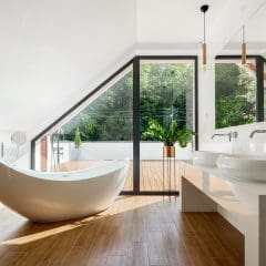 A modern bathroom with a freestanding white bathtub, dual sinks, and a large mirror. Floor-to-ceiling windows offer a view of greenery outside. Light wood flooring and minimalist decor create an airy feel. Towels are placed neatly on a wooden stand.