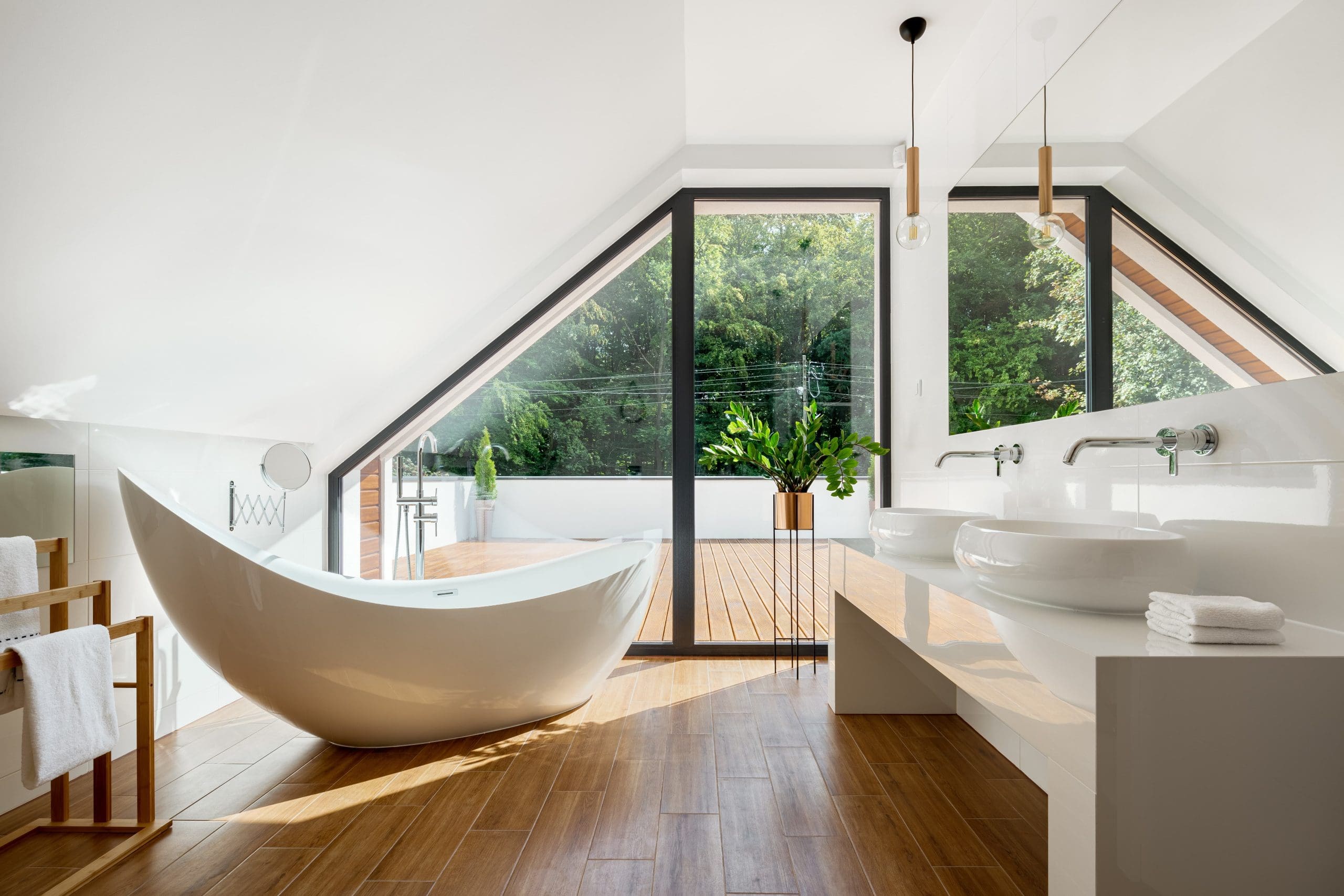 A modern bathroom with a freestanding white bathtub, dual sinks, and a large mirror. Floor-to-ceiling windows offer a view of greenery outside. Light wood flooring and minimalist decor create an airy feel. Towels are placed neatly on a wooden stand.
