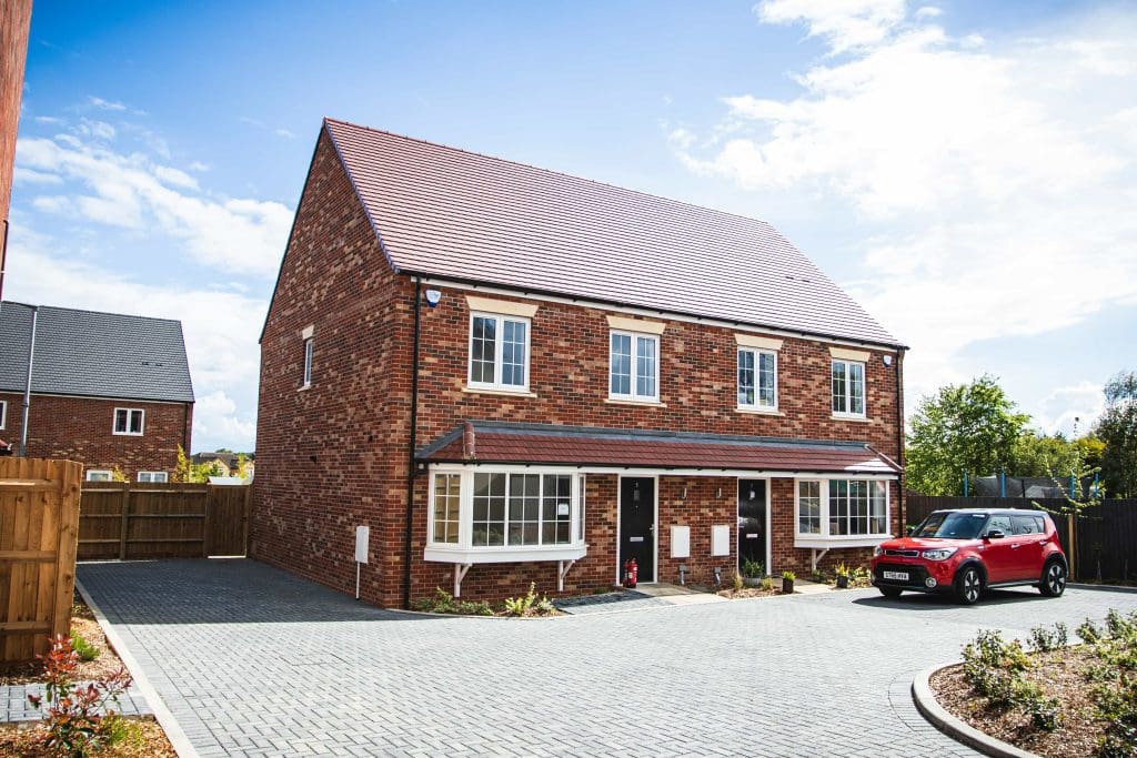 A brick semi-detached house with two entrances and small gardens in front, located on a paved driveway. A red car is parked to the right. The sky is clear and sunny, with other similar houses visible in the background.